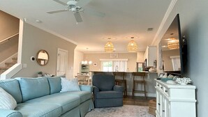 A living room with a light blue sofa, a gray armchair, and a patterned rug. There is a ceiling fan, pendant lights, and a television on a white cabinet. - Beach Getaway: Stunning, Family-Friendly w/ Pool. Walk to Shopping + Dining (Atlantic Beach)
