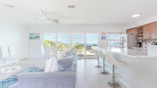 A modern living room with a grey sofa, white chairs, and a kitchen area with wooden cabinets and a stainless steel refrigerator.
