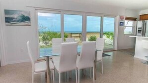A dining area with a glass table and white chairs, overlooking a beach and ocean view. - Oceanfront Modern Home w/ Private Beach (Brevard County)