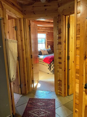 L’intérieur d’un chalet en bois présente une chambre visible, avec un couvre-lit et des oreillers à carreaux, une fenêtre laissant entrer la lumière du jour et un couloir menant à une autre pièce.
