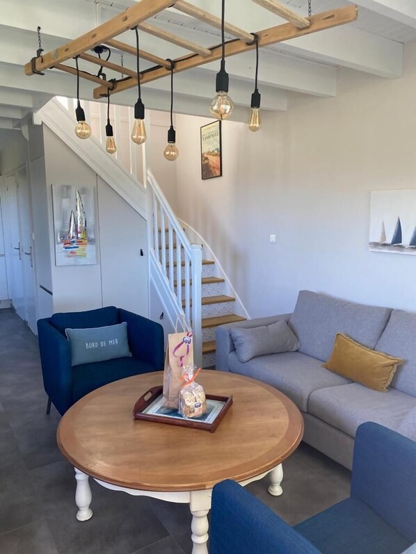 A modern living room with a wooden coffee table, blue and grey sofas, a staircase, and hanging light fixtures. - Maison Bord de mer (Landunvez)