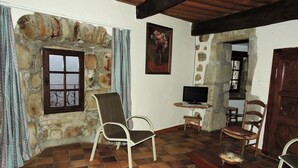 Una habitación rústica con paredes de piedra, piso de baldosas, un televisor sobre un soporte de madera y dos sillas.