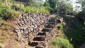 Una escalera de piedra con barandales de metal que asciende por una ladera cubierta de césped, rodeada de árboles y vegetación.