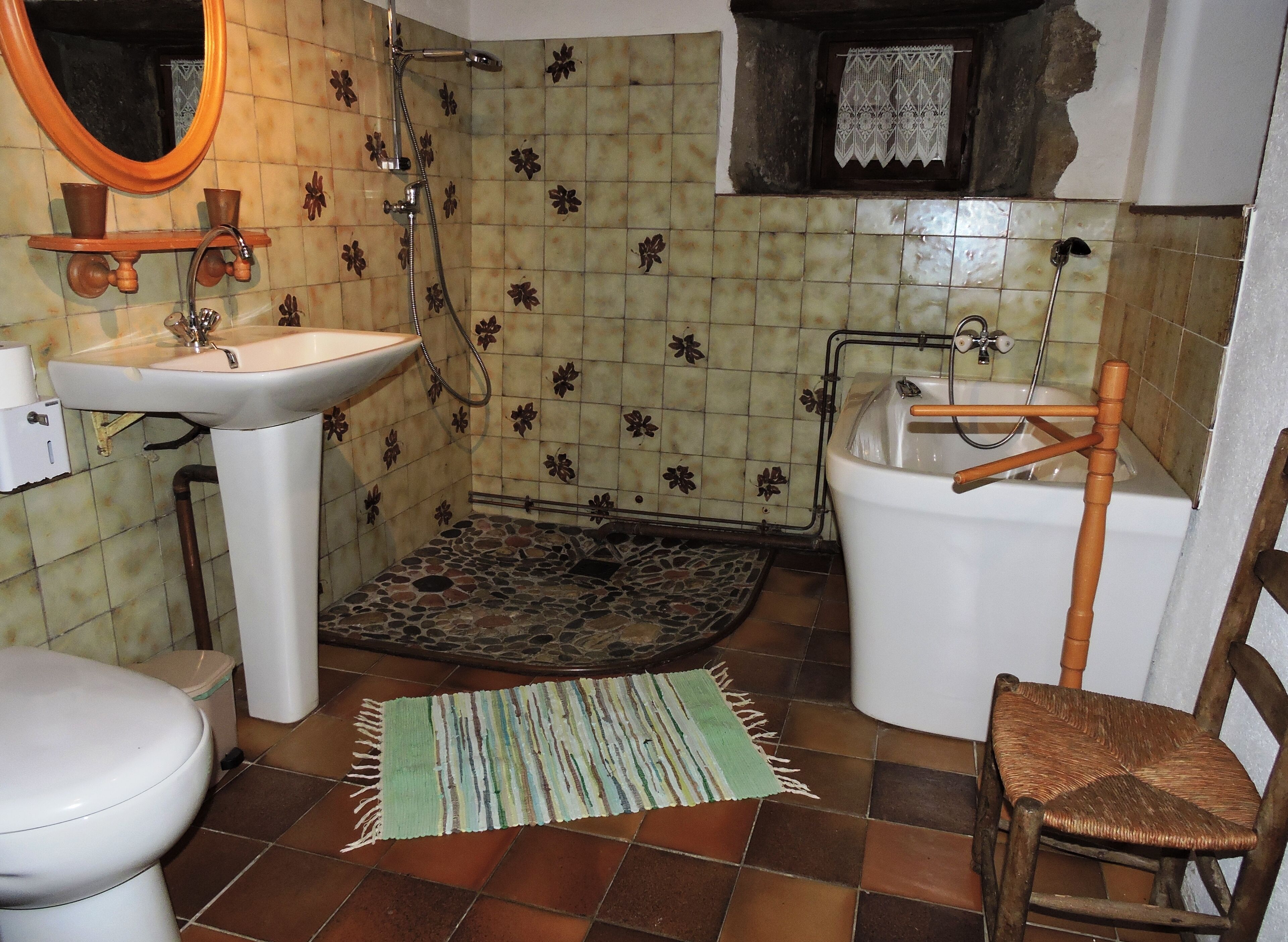A bathroom with a white sink, a toilet, a shower with a wooden chair, and a patterned floor.