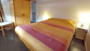 A bedroom with a bed, a chair, a wardrobe, and a bedside table with a lamp. - Maisonnette Biscarrosse Plage 300m de Locéan (Biscarrosse)