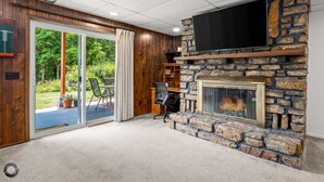 A room with a stone fireplace, a flat-screen TV mounted above it, and a sliding glass door leading to an outdoor area with a patio and greenery. - Lakefront Home w/ Private Dock & MTB Trails Minutes from Bentonville (Bella Vista)