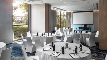 Un salón de conferencias con mesas redondas, sillas blancas y una pantalla para proyector.