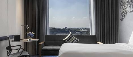 Una habitación de hotel con cama, escritorio, silla, una mesita con lámpara y vistas a la ciudad a través de grandes ventanales.