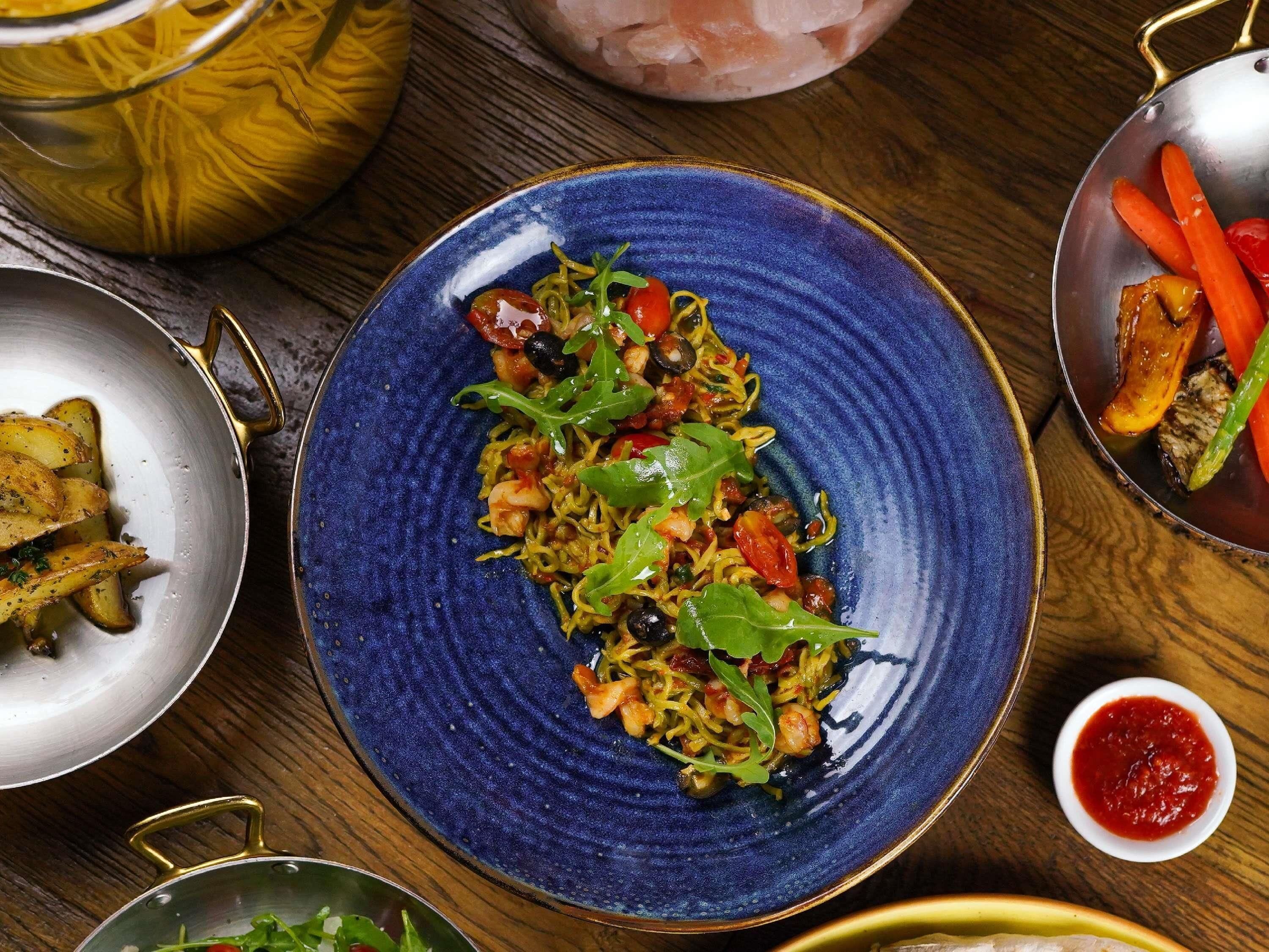 A blue plate with a salad, a bowl of sauce, and a pot of food on a wooden table.