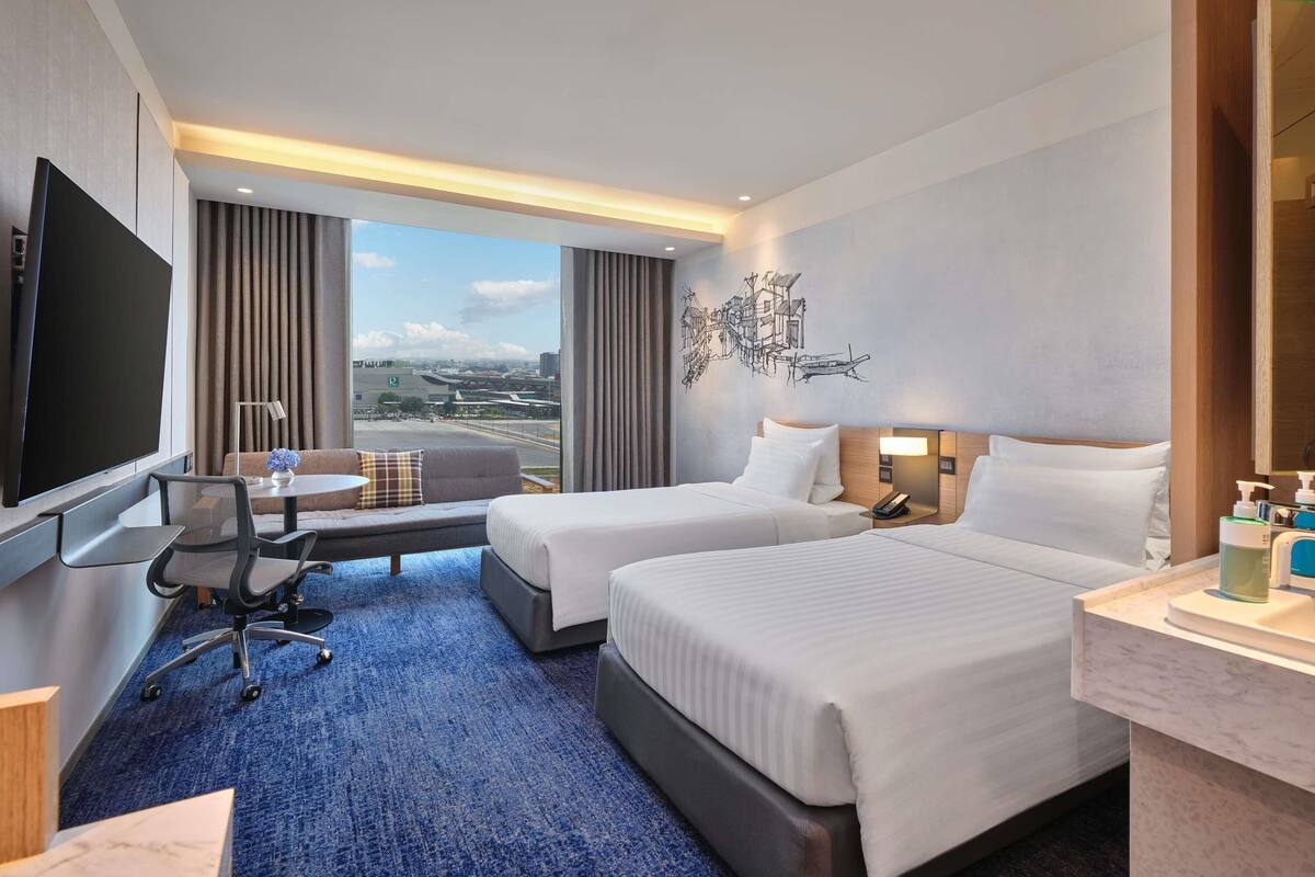 A hotel room with two beds, a desk, a chair, a TV, and a large window with a view of an airport.