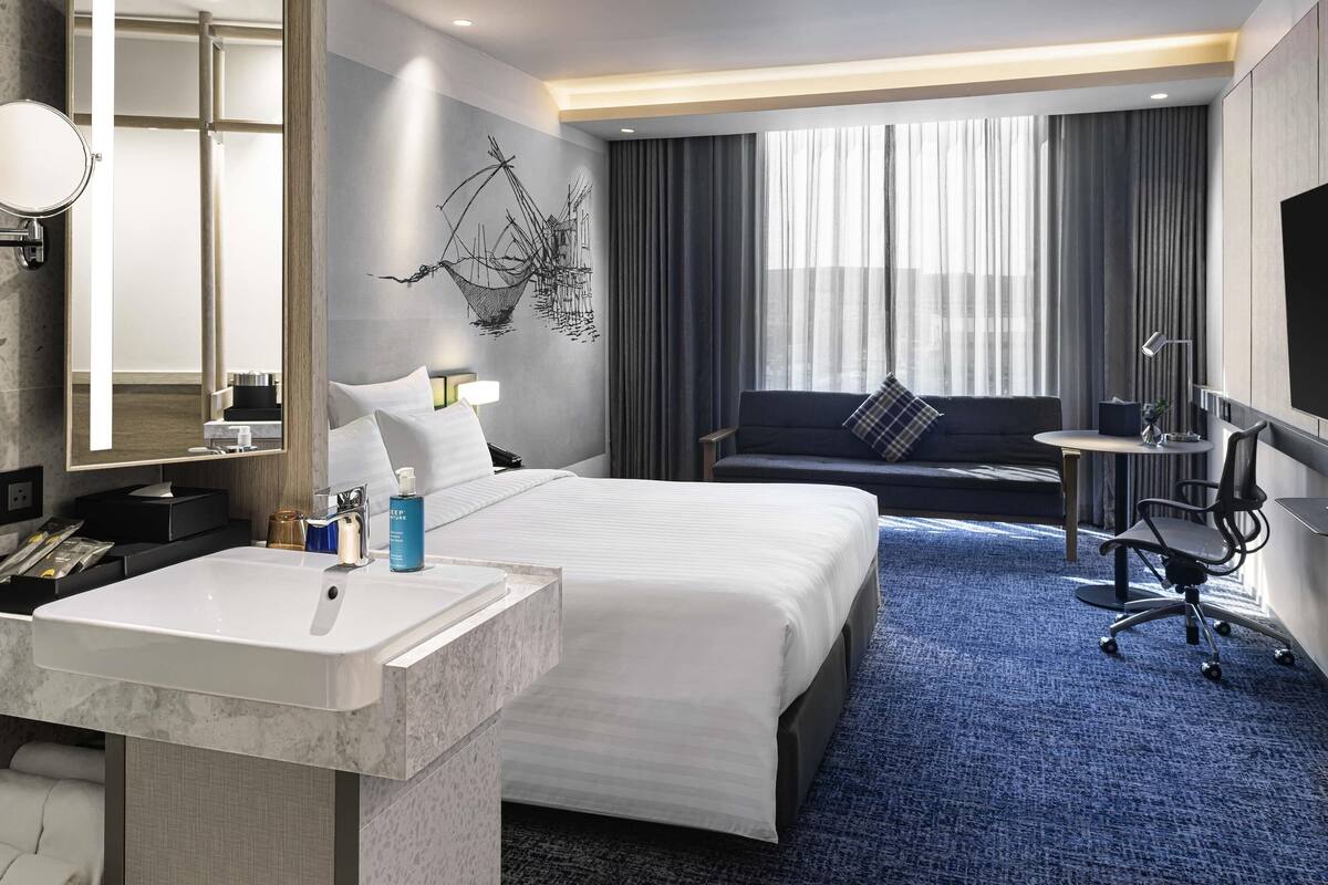 A hotel room with a large bed, a sitting area with a sofa and armchair, a desk with a chair, a large window with curtains, a wall-mounted TV, and a bathroom with a sink and mirror.