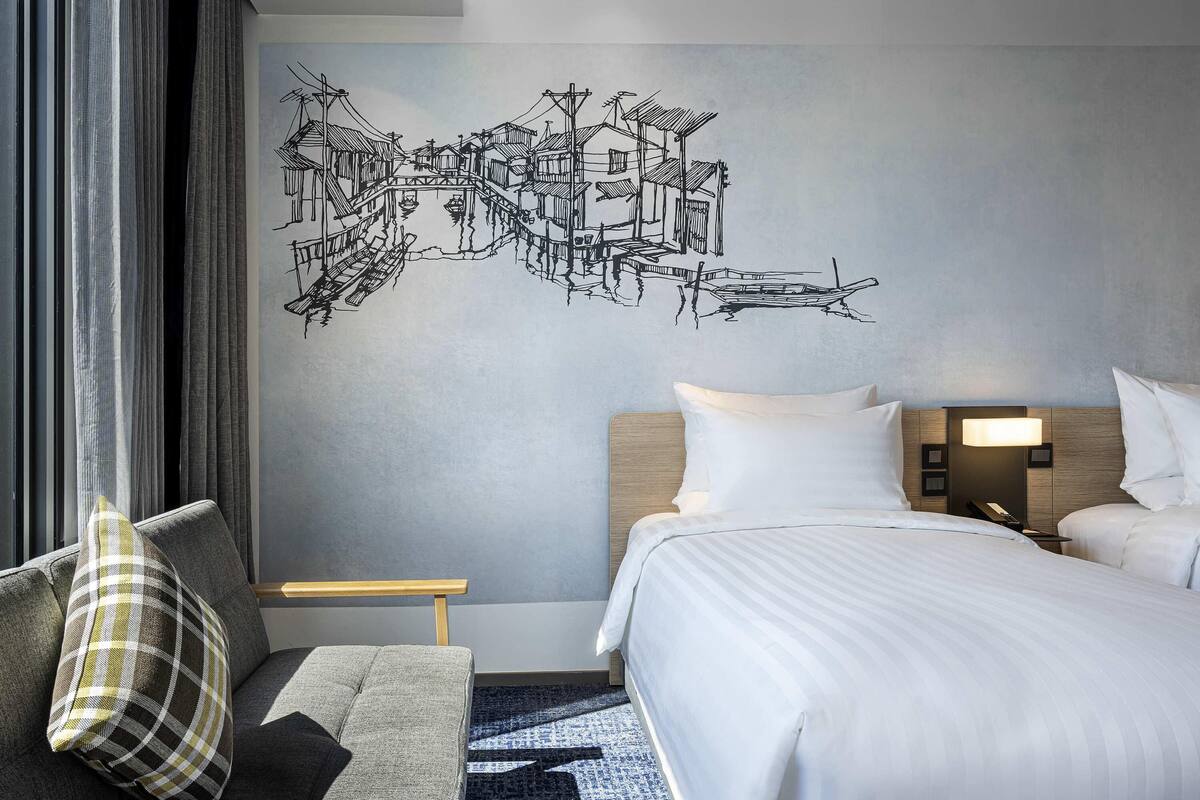 A hotel room with a bed, a nightstand, a lamp, a sofa with a pillow, and a wall with a black and white mural.