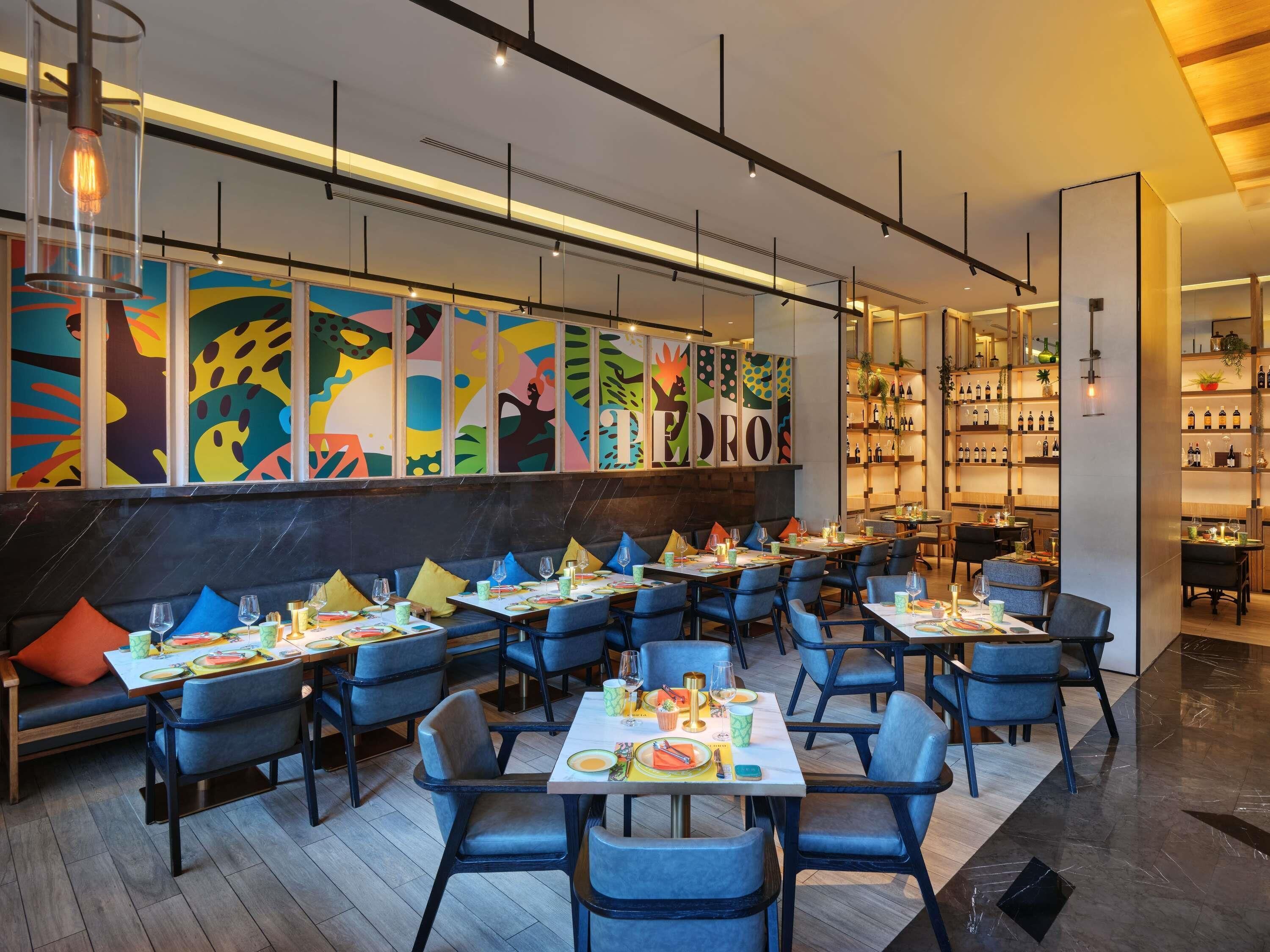 Un restaurante moderno con varias mesas y sillas, arte abstracto colorido en la pared y estantes con botellas.