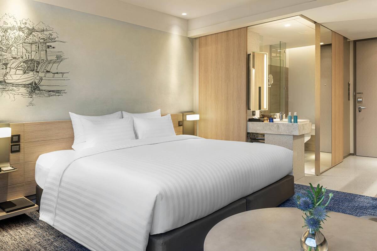 A modern hotel room with a large bed, bedside tables, a nightstand, a round coffee table, and a bathroom with a mirror and sink.