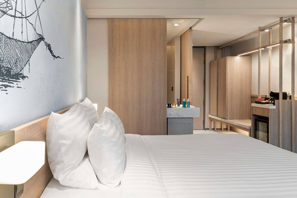A modern hotel room with a large bed, a bedside table, a mirror, a vanity with a sink, and a wardrobe.