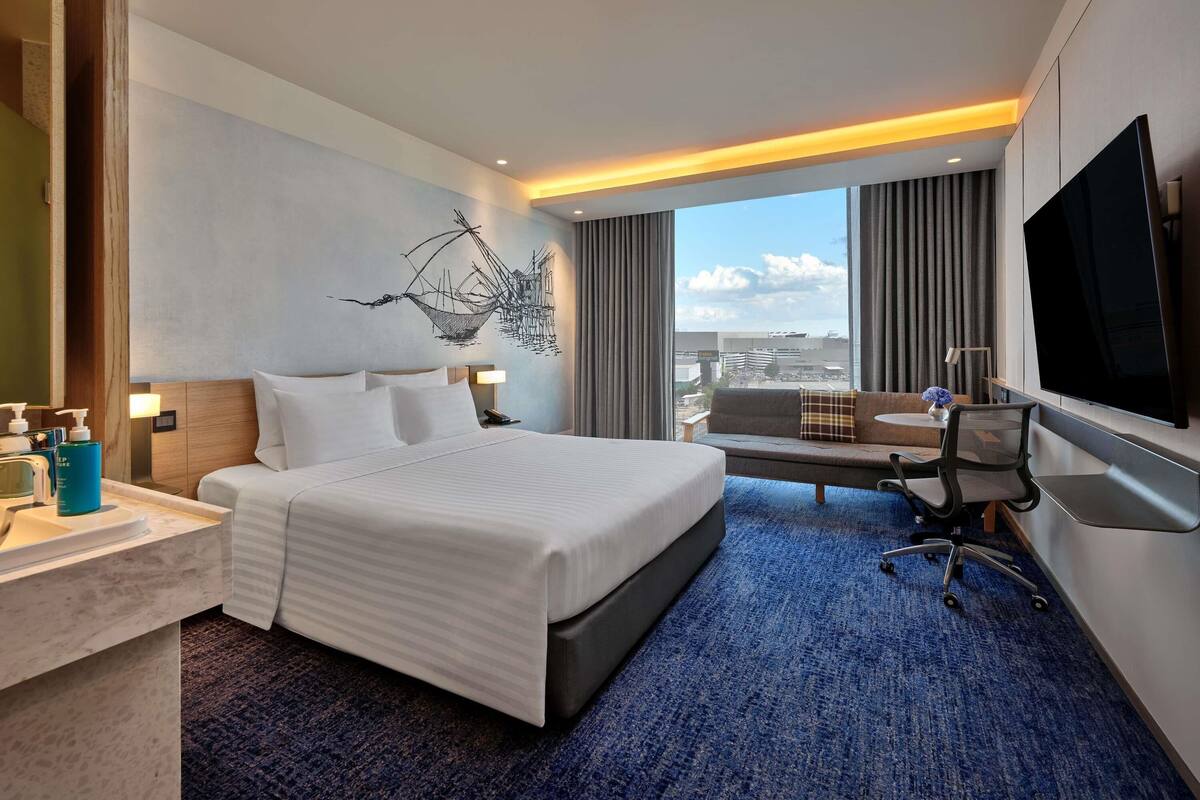 A hotel room with a bed, a desk, a chair, a TV, and a large window with a city view.