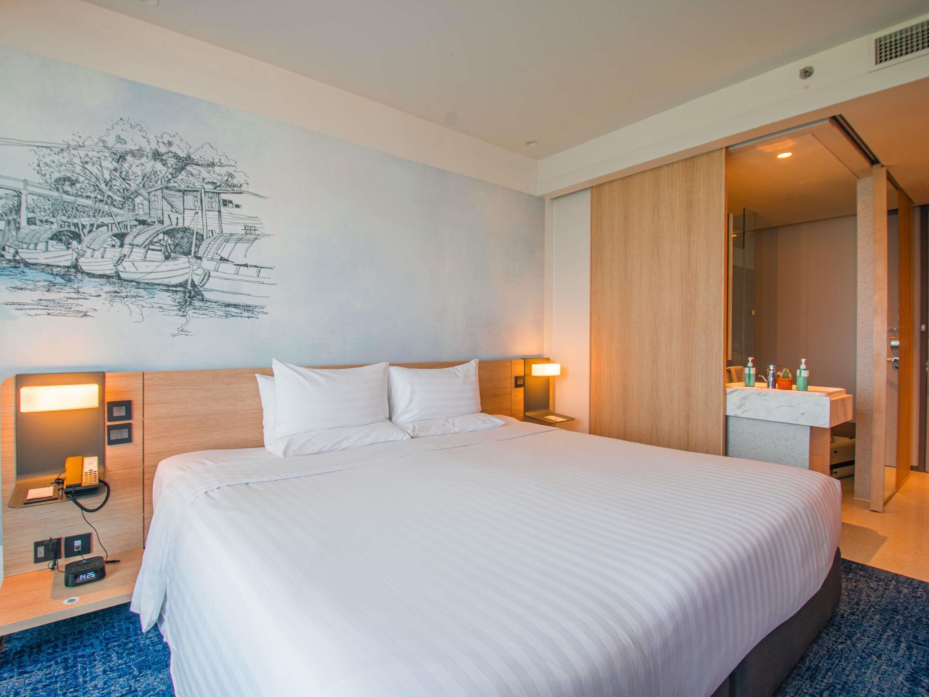 Habitación de hotel con una cama grande, lámparas de noche, mesita de noche, un mural en la pared y un baño visible a través de una puerta abierta.