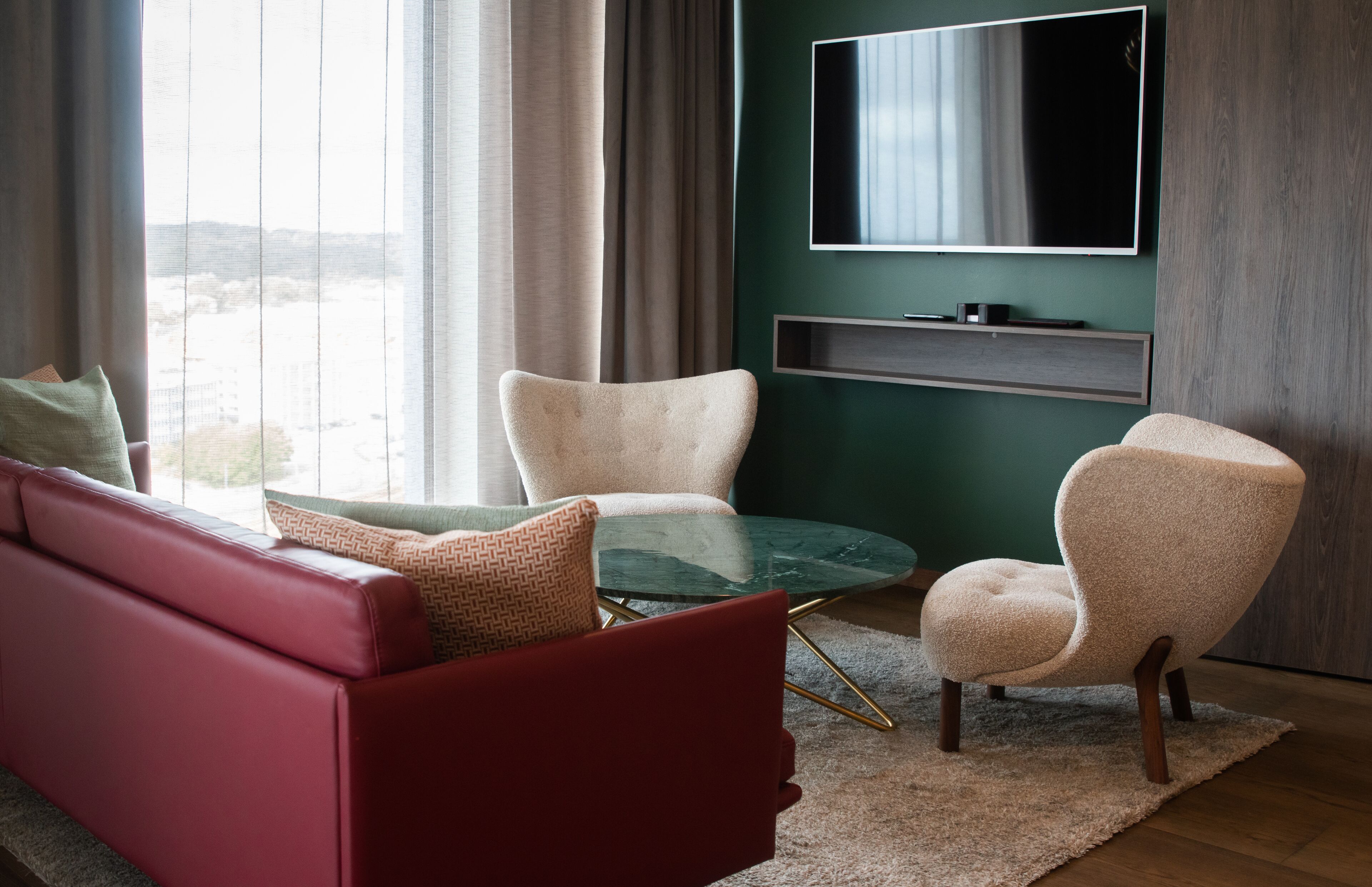 A modern living room with a red sofa, two armchairs, a glass coffee table, and a flat-screen TV mounted on a green wall.
