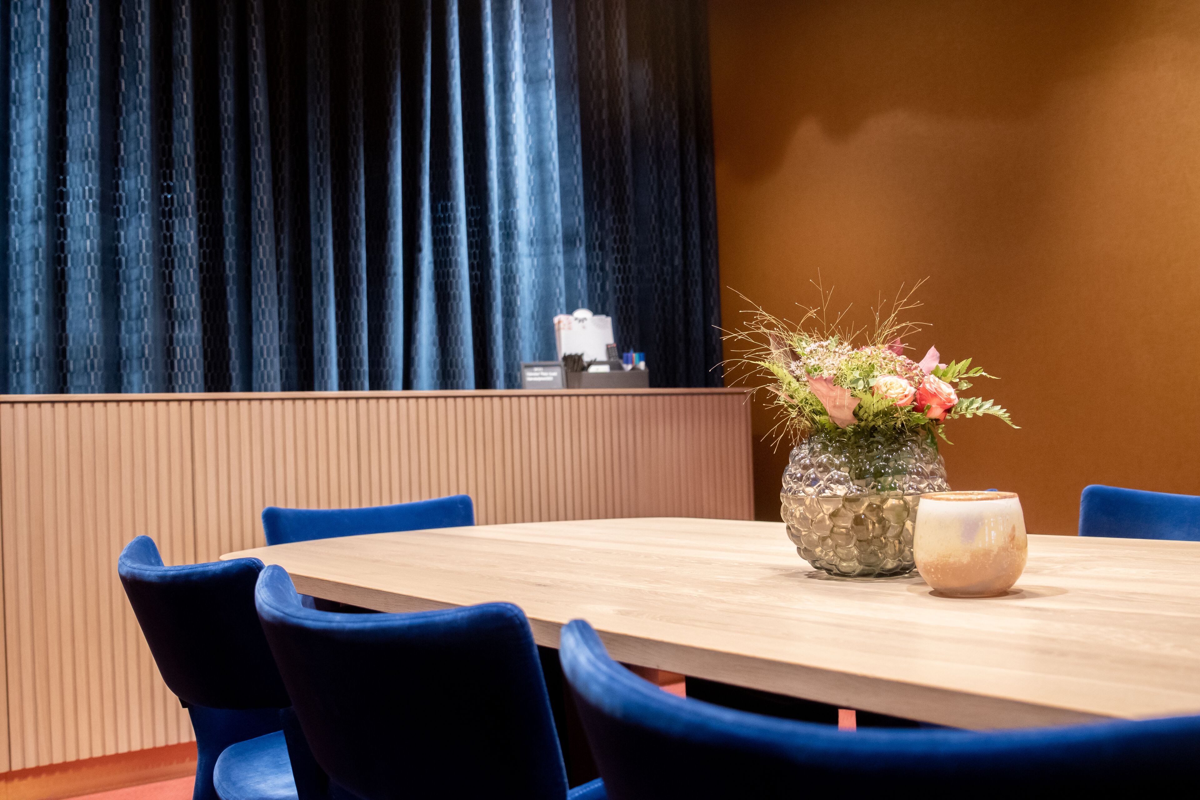 A conference room with a wooden table, blue chairs, a floral centerpiece, and a coffee pot.