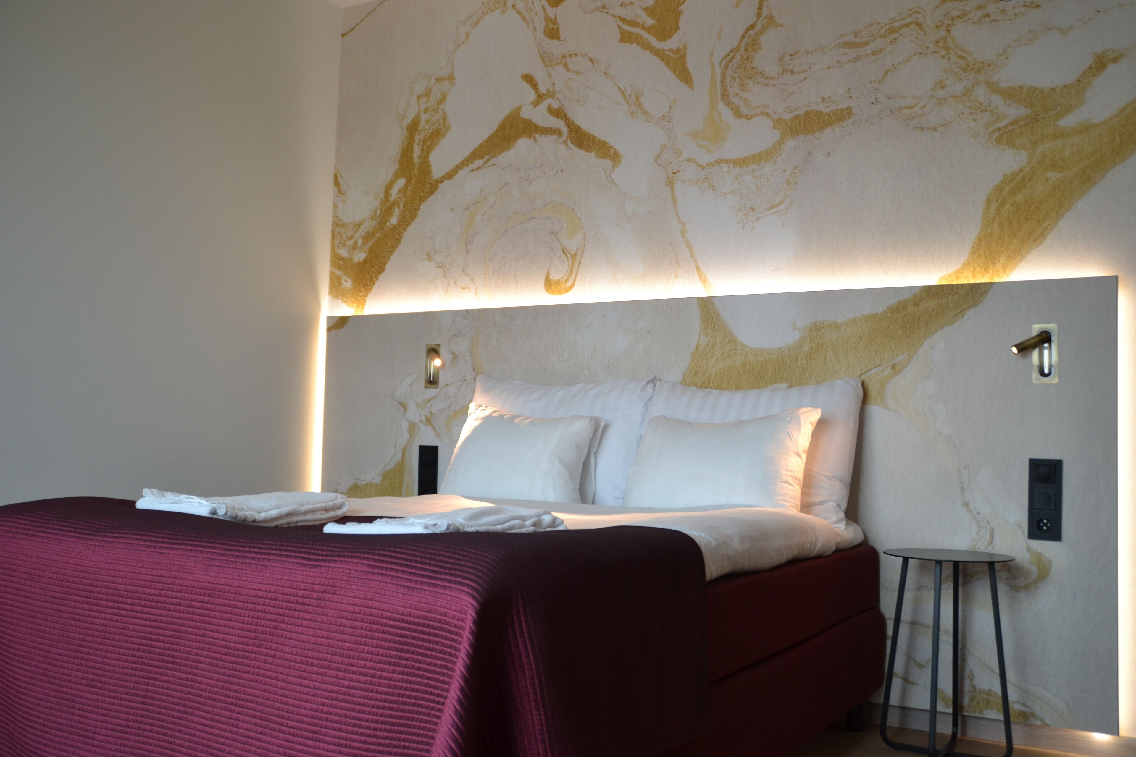 Habitación de hotel con una cama grande, una mesita auxiliar pequeña y un muro con diseño de mármol.