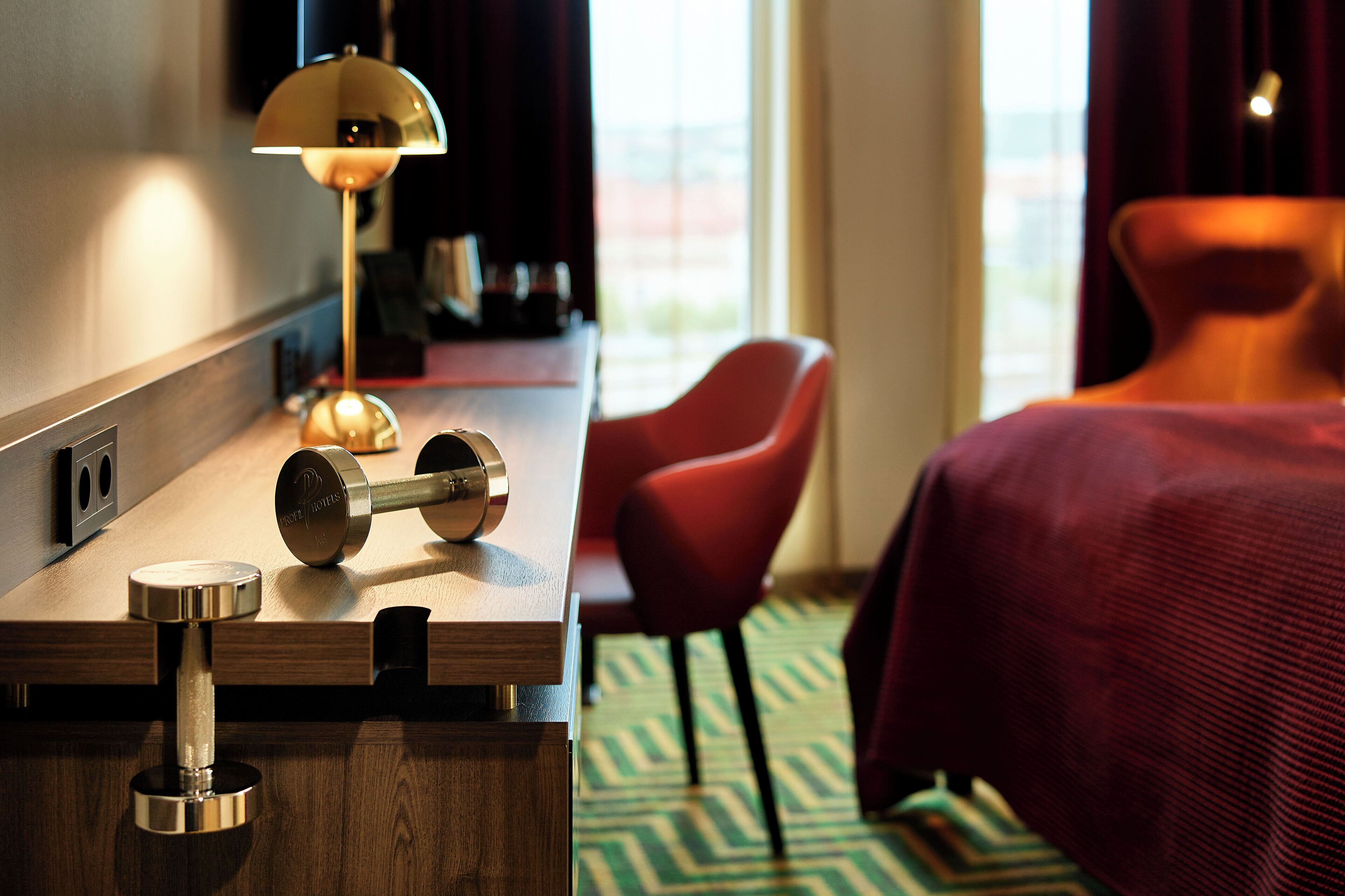 Una habitación de hotel con un escritorio de madera, una lámpara, una silla roja y una cama con cubrecolcha roja.