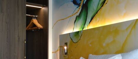 A hotel room with a bed, a wooden wardrobe, and a wall with a colorful abstract painting.