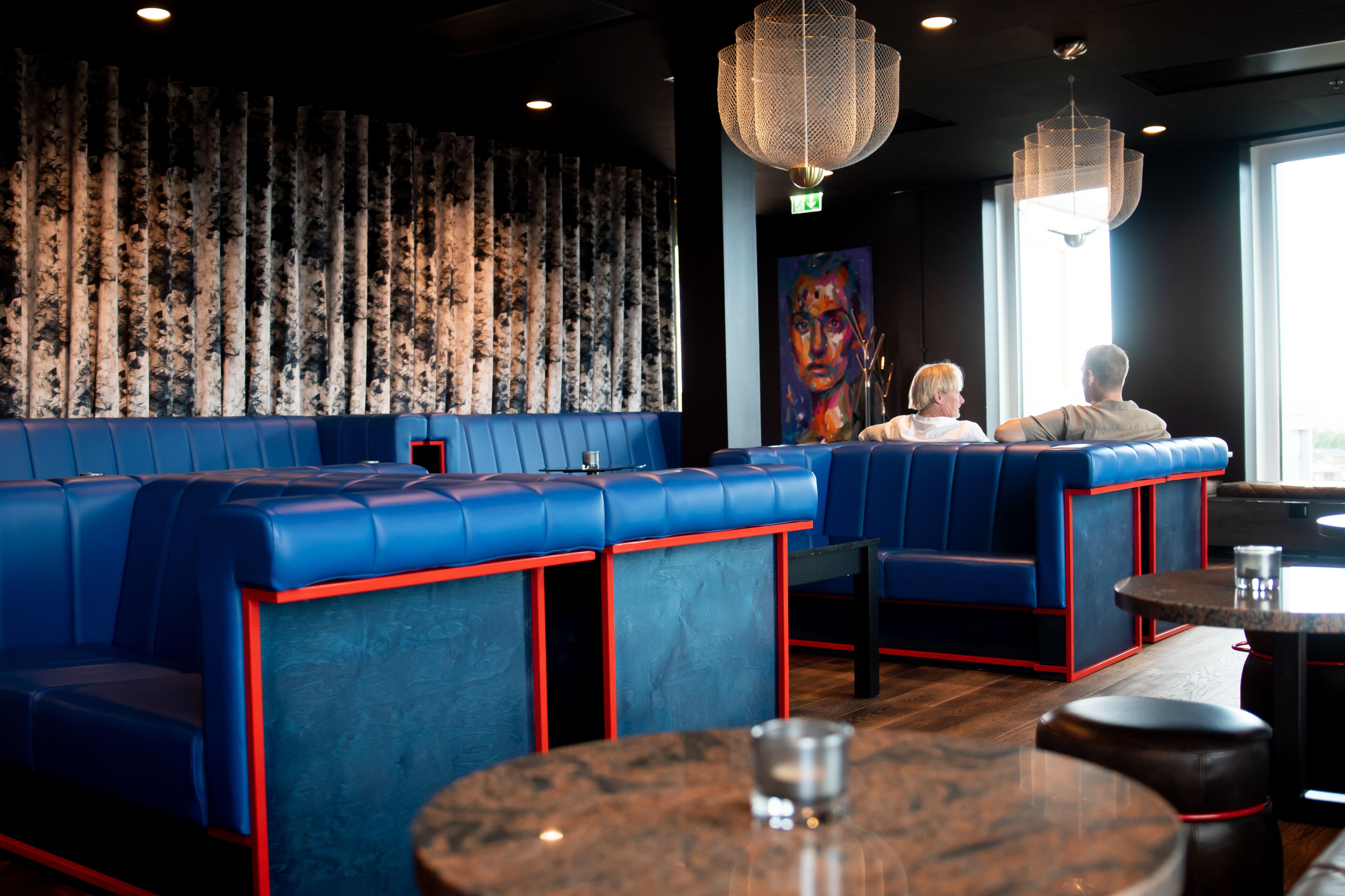 En restaurang med blå bås och röda detaljer, en stor målning på väggen och en kristallkrona.