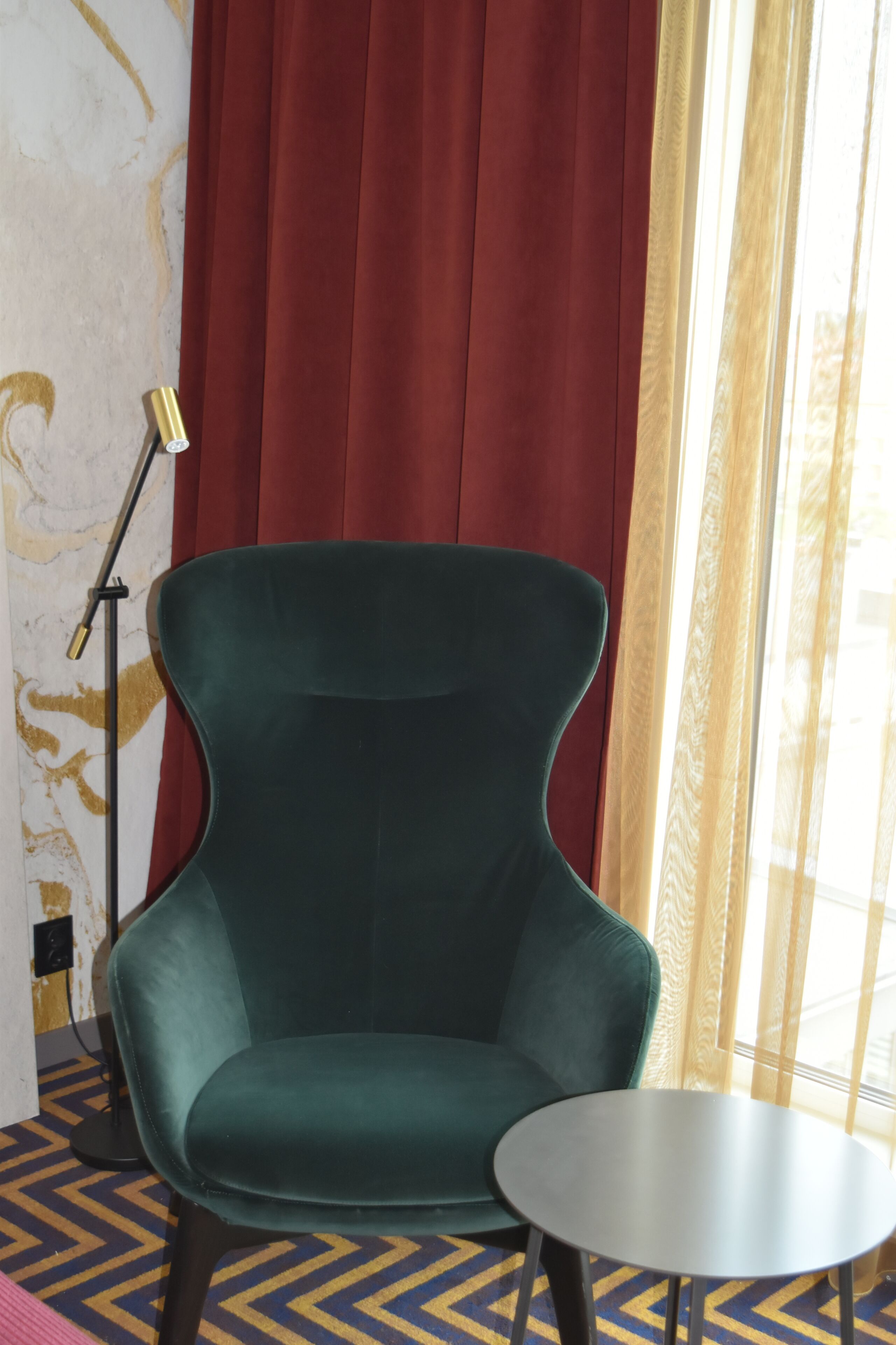 A room with a green armchair, a small round table, a floor lamp, and a window with curtains.