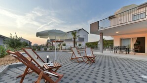 A patio area with wooden lounge chairs and a table, a large umbrella, and a building with a balcony. - Exquisite 4 BD villa for 8 with heated pool near Zadar (Sukošan)