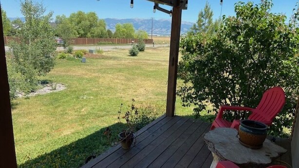 Een overdekt terras met rode stoelen en een tafel, met uitzicht op een gazon met bomen en bergen op de achtergrond.