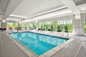 An indoor swimming pool with clear views of trees through large windows, surrounded by lounge chairs and a tiled floor.