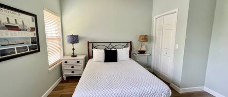 Un dormitorio con cama, ventilador de techo, mesita de noche con lámpara, un cuadro enmarcado en la pared y una puerta.