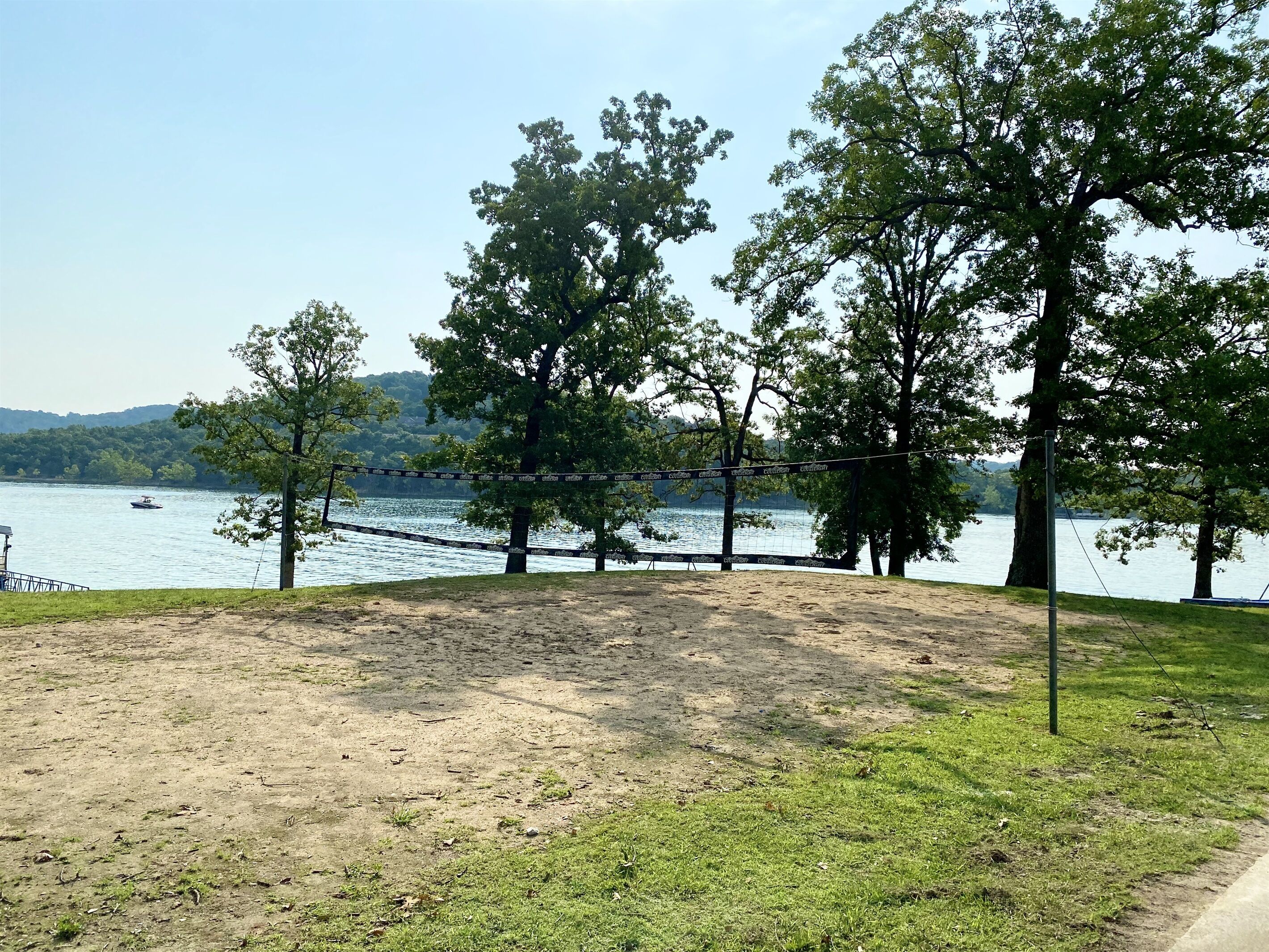 Ein Sandvolleyballfeld mit Netz, umgeben von Bäumen und einem See mit Booten im Hintergrund.