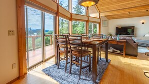 Uma sala de jantar espaçosa com mesa e cadeiras de madeira, grandes janelas com vista para o exterior e teto com vigas de madeira.