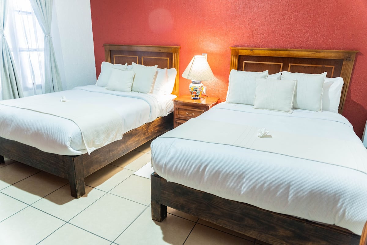 Two single beds with wooden frames and white bedding, a nightstand with a lamp, and a red wall.