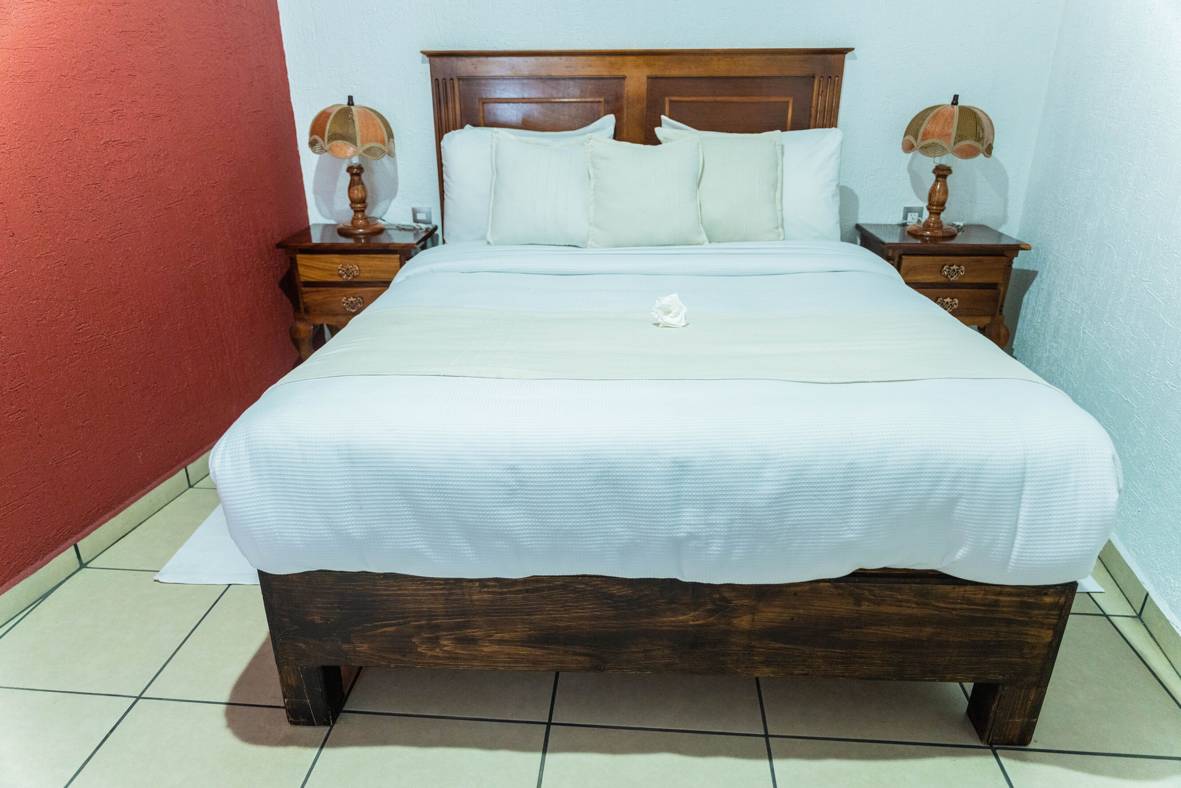 Una cama bien tendida con un cabecero de madera, dos mesitas de noche con lámparas y una pared roja.