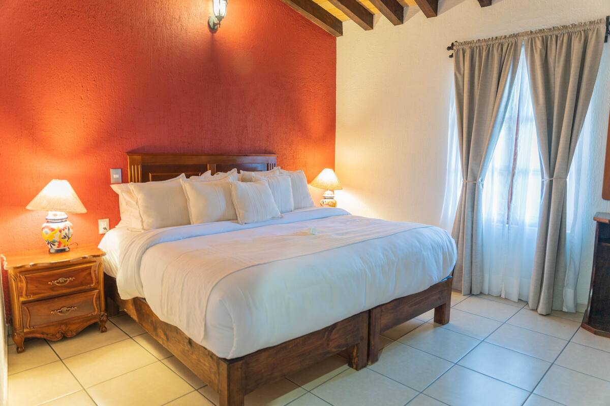 A bedroom with a wooden bed, two bedside tables with lamps, a red wall, and a window with curtains.