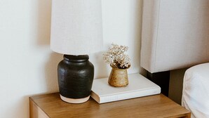 A wooden bedside table with a lamp, a book, and a small vase with dried flowers. - Serene Ocean View, Waterfront Cottage Near Olympic National Pk (Port Angeles)