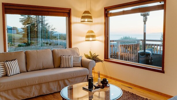 A cozy living room with a sofa, a glass coffee table, and two large windows offering a view of the outdoors. - Serene Ocean View, Waterfront Cottage Near Olympic National Pk (Port Angeles)
