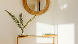 A room with a wooden floor, a round mirror on the wall, a small table with a plant and a book, and a white bed in the corner. - Serene Ocean View, Waterfront Cottage Near Olympic National Pk (Port Angeles)