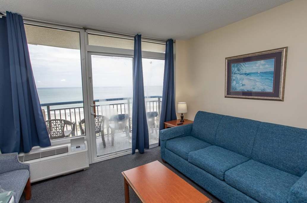 Habitació amb un sofà blau, una taula de fusta, una cadira, una làmpada i vista a l'oceà a través d'una porta de vidre.