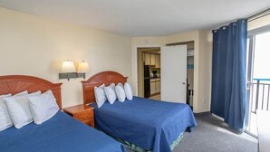 A hotel room with two beds, a nightstand, a lamp, a door leading to a kitchen, and a balcony with curtains. - Boardwalk 539 & 540 (Myrtle Beach)