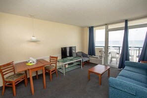 Habitación de hotel con mesa de comedor, sillas, sofá, televisor y balcón con cortinas.