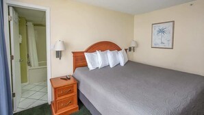 Quarto de hotel com cama, luminárias de cabeceira, mesa de cabeceira de madeira, um quadro pendurado na parede e um banheiro visível através de uma porta aberta.