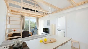 A modern kitchen with a central island, wooden beams, a dining area with a table and chairs, and a view of greenery outside.