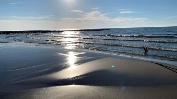 A beach with a pier extending into the water, waves gently lapping at the shore, and the sun reflecting off the wet sand.