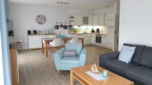 Een moderne keuken met witte kastjes, een eethoek met een houten tafel en stoelen, en een woonkamer met een grijze bank en een blauwe fauteuil.