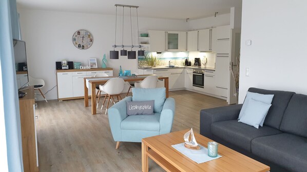 A modern kitchen with white cabinets, a dining area with a wooden table and chairs, and a living room with a grey sofa and a blue armchair.