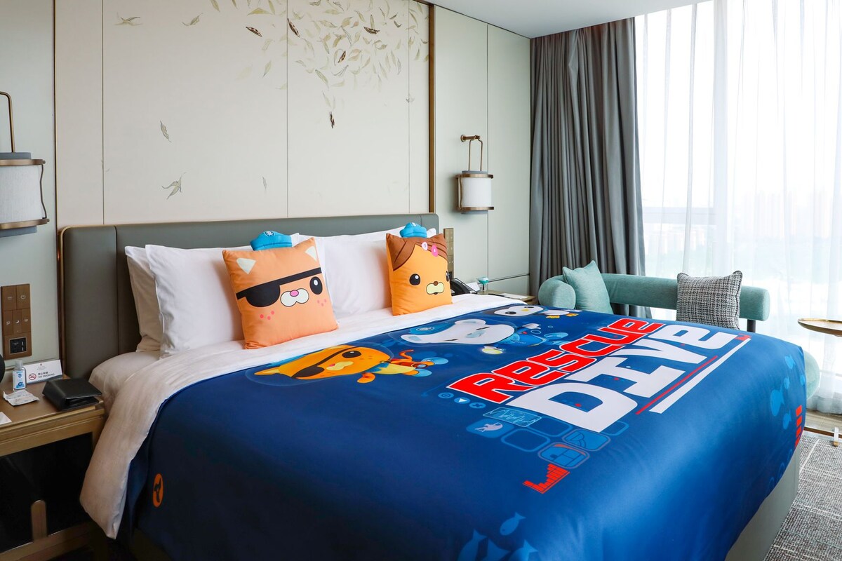 A hotel room with a bed featuring a blue blanket with a Rescue Dogs International logo, two pillows with animal designs, a nightstand with a phone and a lamp, and a view of the outdoors through a window with curtains.