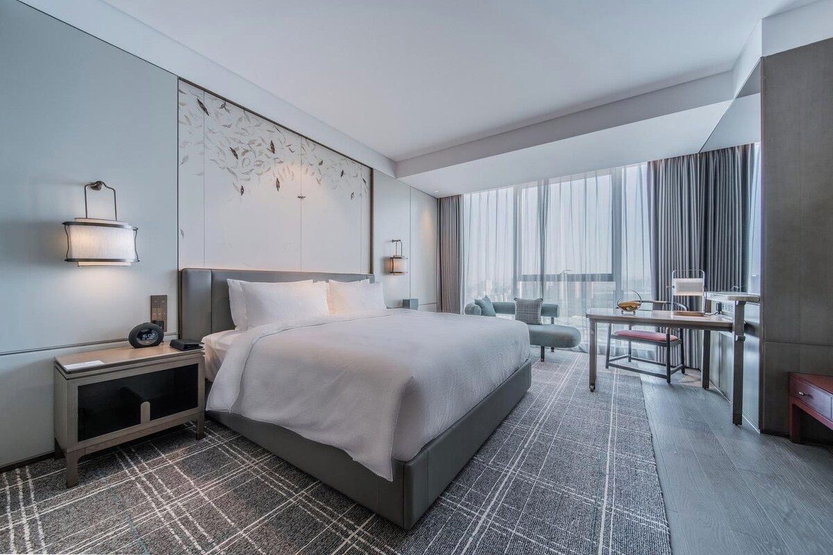 A modern hotel room with a large bed, a desk, and a view of the city through large windows.